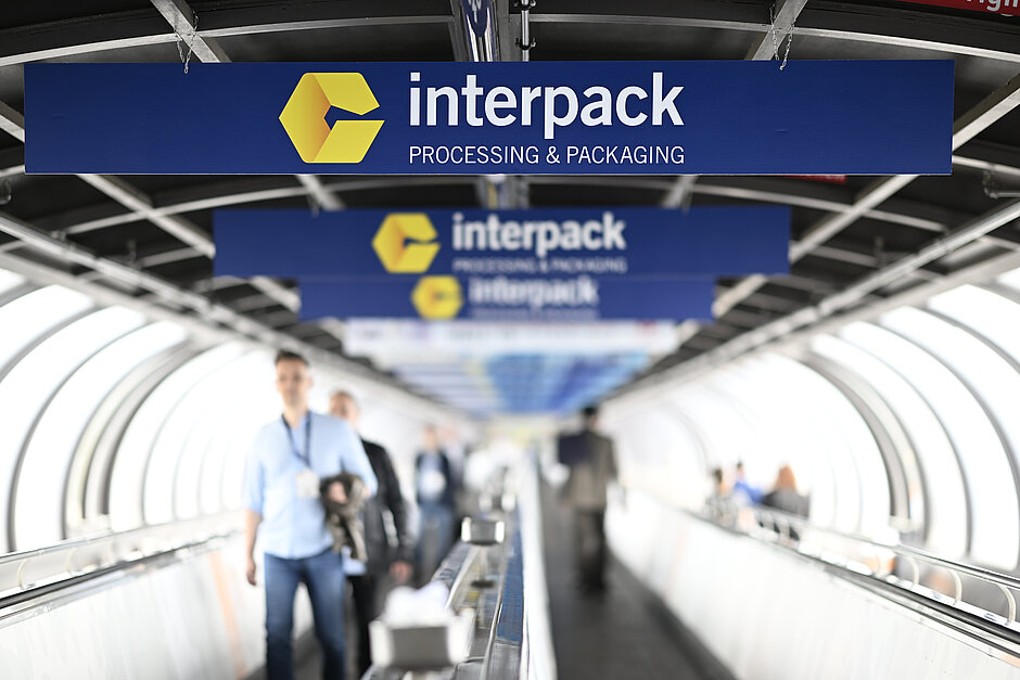 Copyright: Constanze Tillmann, Mintarder Weg 34, 40472 Duesseldorf, Germany People walking along a conveyor belt beneath several blue signs featuring the yellow interpack logo and the text “interpack PROCESSING & PACKAGING” in a tunnel-like structure.